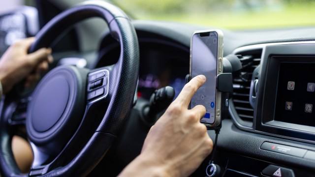 A driver uses a smartphone mounted on the dashboard while gripping the steering wheel.