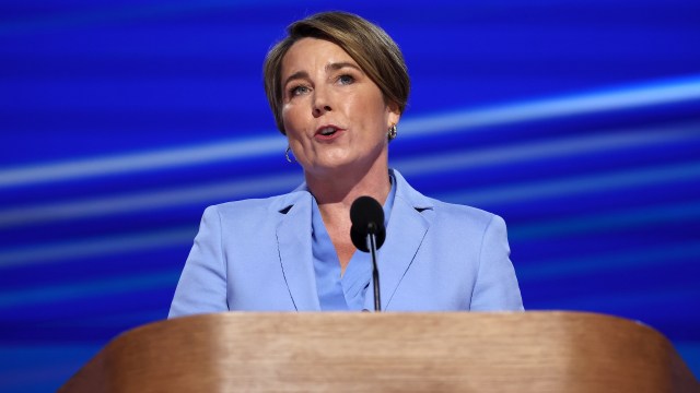 Gov. Maura Healey speaks at a podium with a microphone, set against a vibrant blue background.