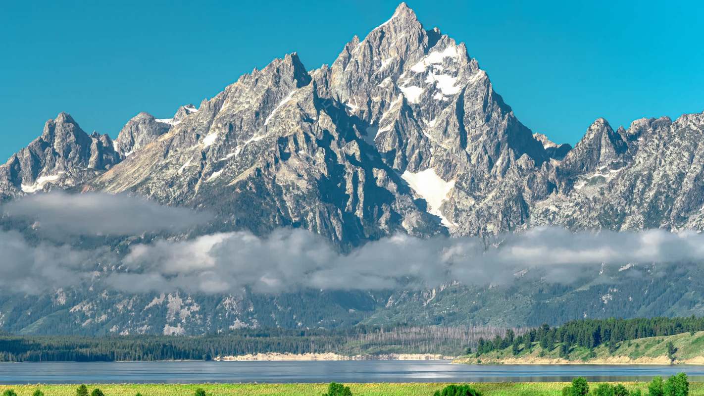 A mountain range with jagged peaks and snow, reflecting in a calm lake under a clear blue sky.