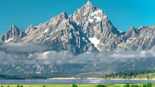 A mountain range with jagged peaks and snow, reflecting in a calm lake under a clear blue sky.