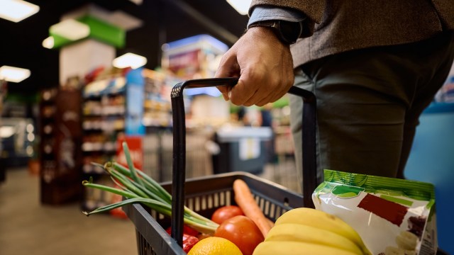 A person holding a shopping basket filled with vegetables and fruits in a grocery store.