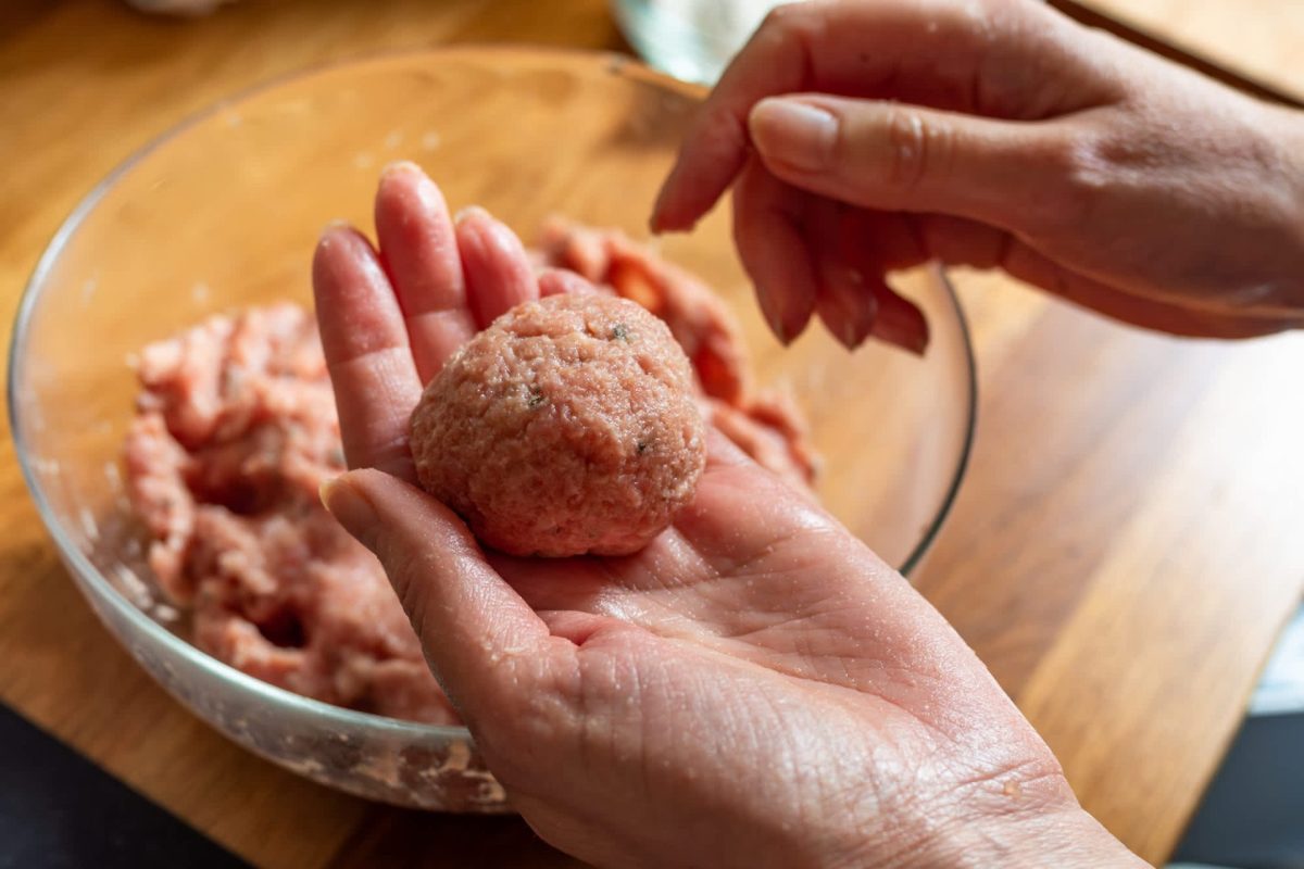 A person forms a meatball in their hand with a bowl of raw ground meat in the background.