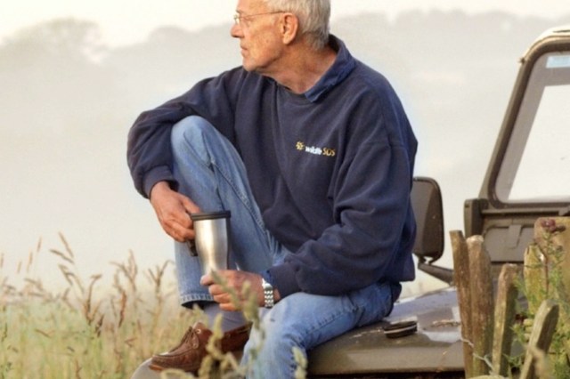 An older man sits on the hood of a Jeep and holds a travel mug.