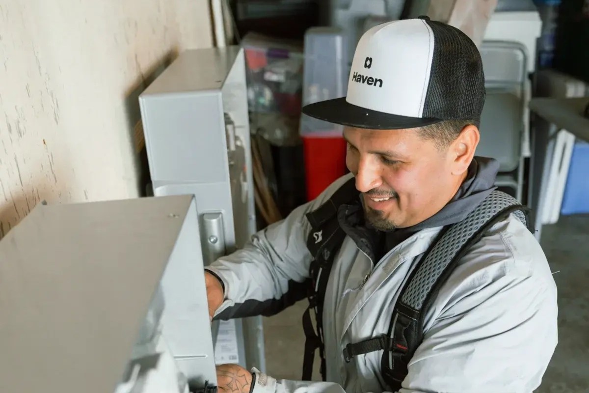 A technician in a gray jacket repairs equipment while smiling in a garage setting.