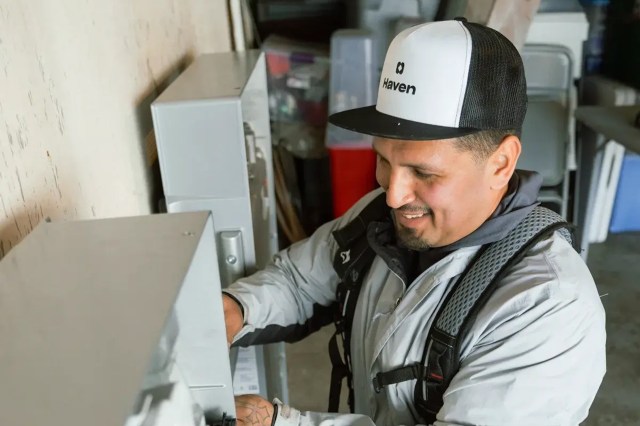 A technician in a gray jacket repairs equipment while smiling in a garage setting.