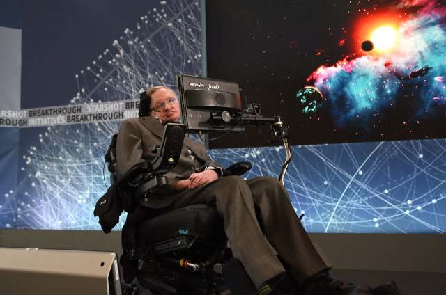 Stephen Hawking, in a wheelchair with a communication device, with a cosmic background displayed behind him.