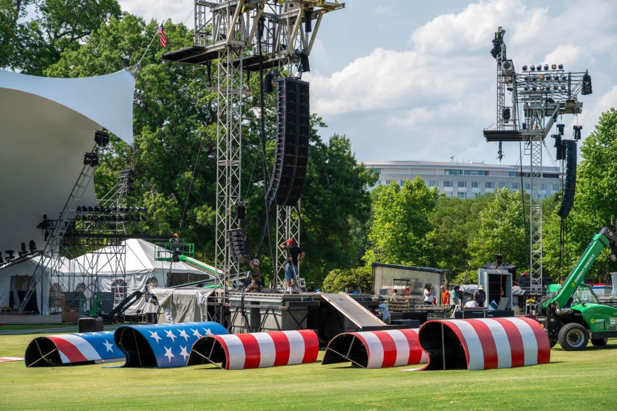 A staging area for an event featuring red, white, and blue structures on grass, with scaffolding and equipment in the background.