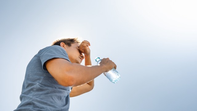 A man holds a water bottle while touching his forehead, with a bright sky in the background.