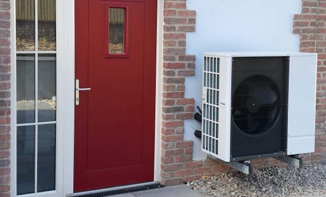 A red front door with a window, next to an outdoor heat pump unit mounted on a brick wall.