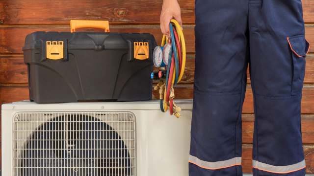 A technician stands holding colorful hoses next to a toolbox on an air conditioning unit.
