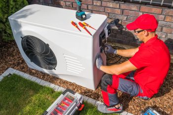 A technician in a red uniform repairs an outdoor HVAC unit using tools on the ground beside him.