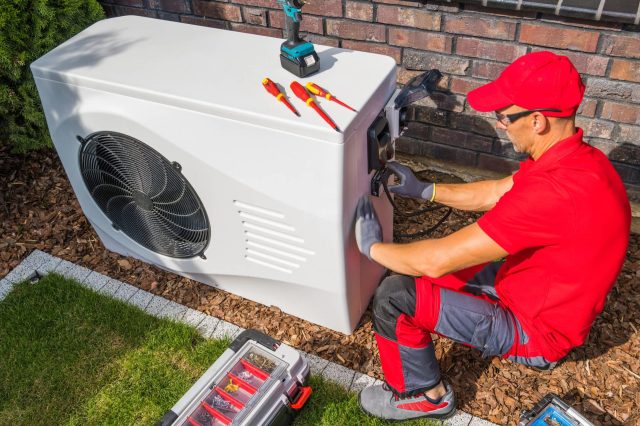 A technician in a red uniform repairs an outdoor HVAC unit using tools on the ground beside him.