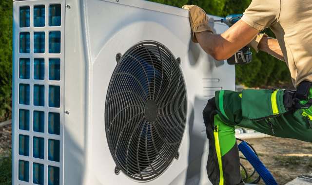 A technician in gloves installs an outdoor heat pump unit with a drill.