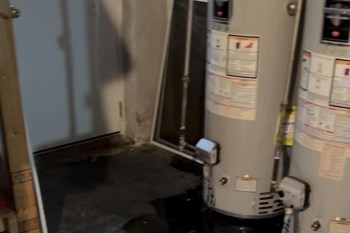 A water heater situated in a basement with a door and some tools visible in the background.