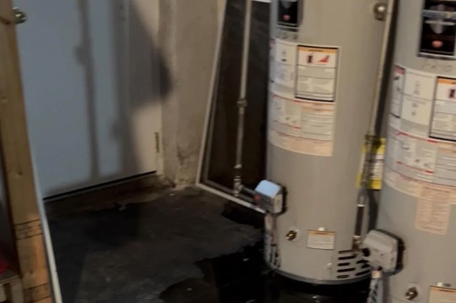A water heater situated in a basement with a door and some tools visible in the background.