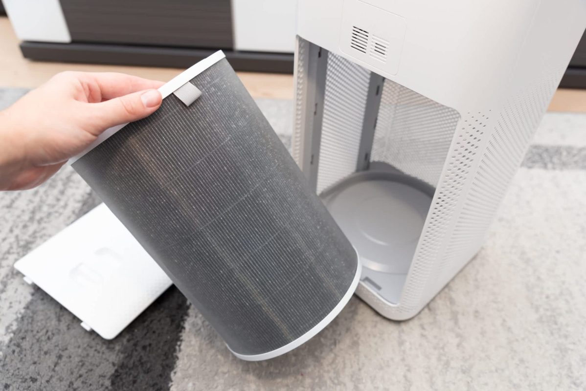 A person holds a dark air purifier filter, preparing to replace it in a white air purifier unit.