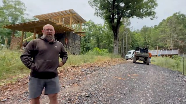 A man stands on a gravel road with his off-grid house and solar panels in a wooded area.