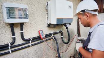 A technician in a hard hat uses a multimeter to test electrical equipment mounted on a wall.