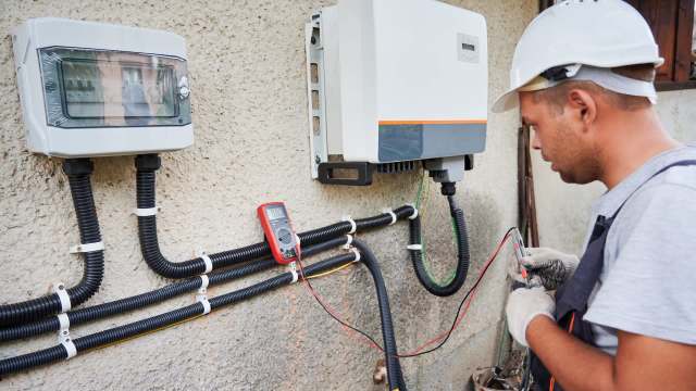 A technician in a hard hat uses a multimeter to test electrical equipment mounted on a wall.