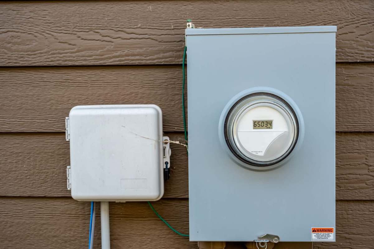 A grey electric meter and a white electrical box mounted on a brown wooden wall.