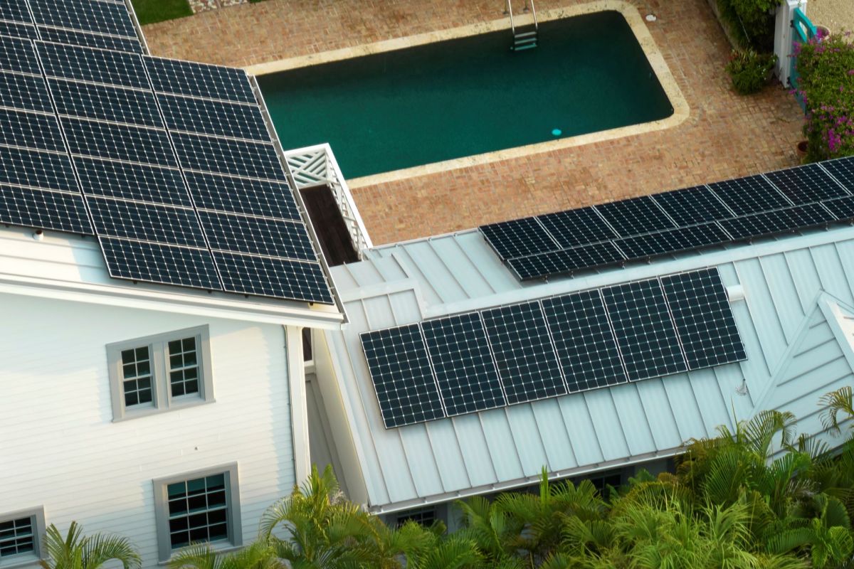 A house with solar panels on the roof, overlooking a swimming pool surrounded by brick patio and greenery.