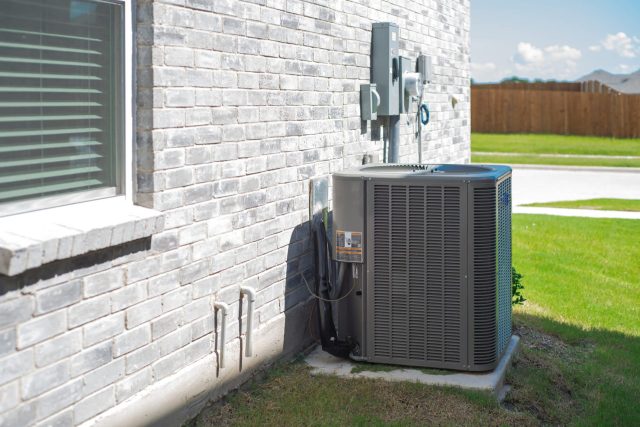 An air conditioning unit is positioned beside a gray brick house with power connections visible on the wall.