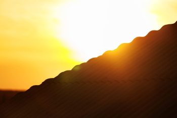 A sun sets behind a wavy rooftop, casting warm orange and yellow hues in the sky.