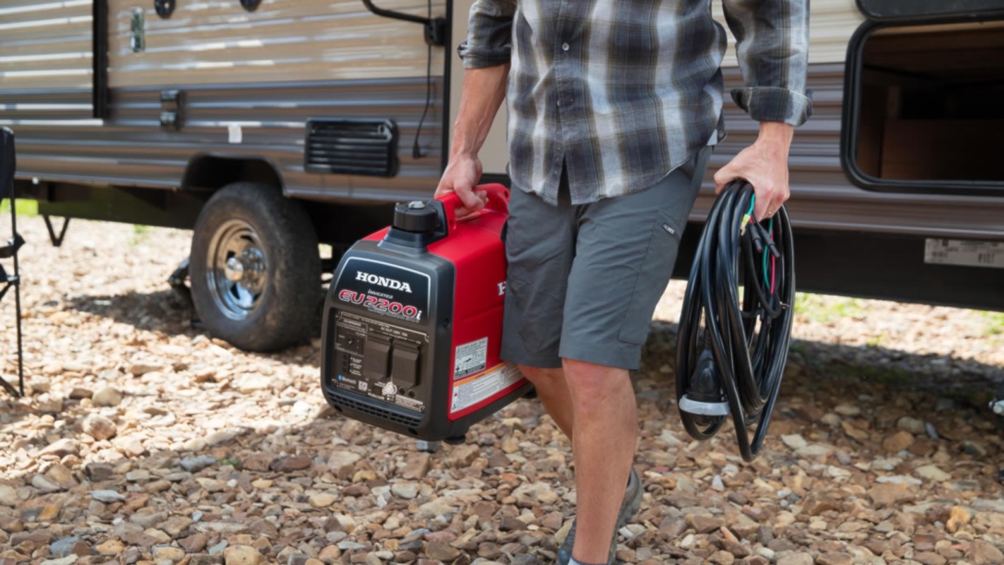 A person carries a Honda generator and electrical cords near a camper in a rocky outdoor setting.