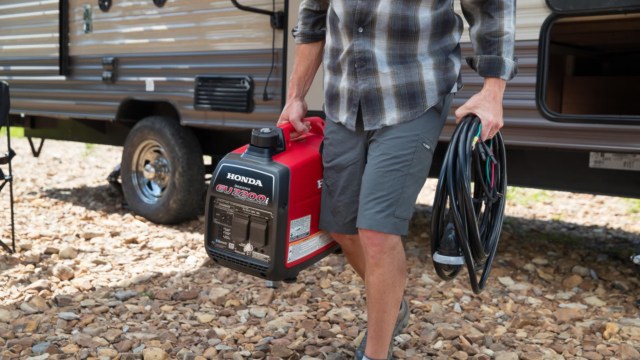 A person carries a Honda generator and electrical cords near a camper in a rocky outdoor setting.