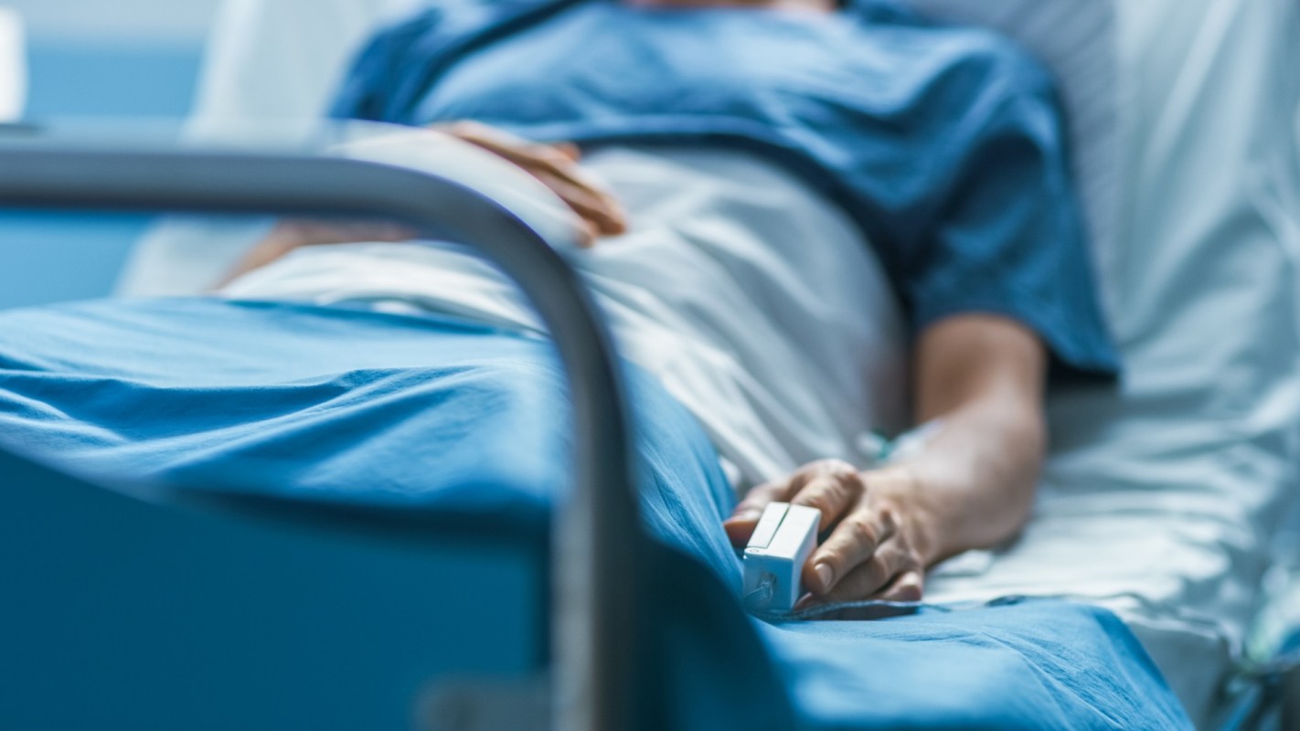 A patient lies in a hospital bed, holding a medical device with monitors in a clinical setting.