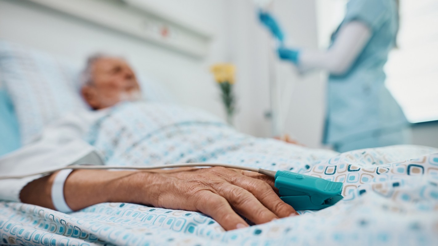 An elderly patient rests in a hospital bed, with a medical monitor attached to their finger and a nurse in the background.