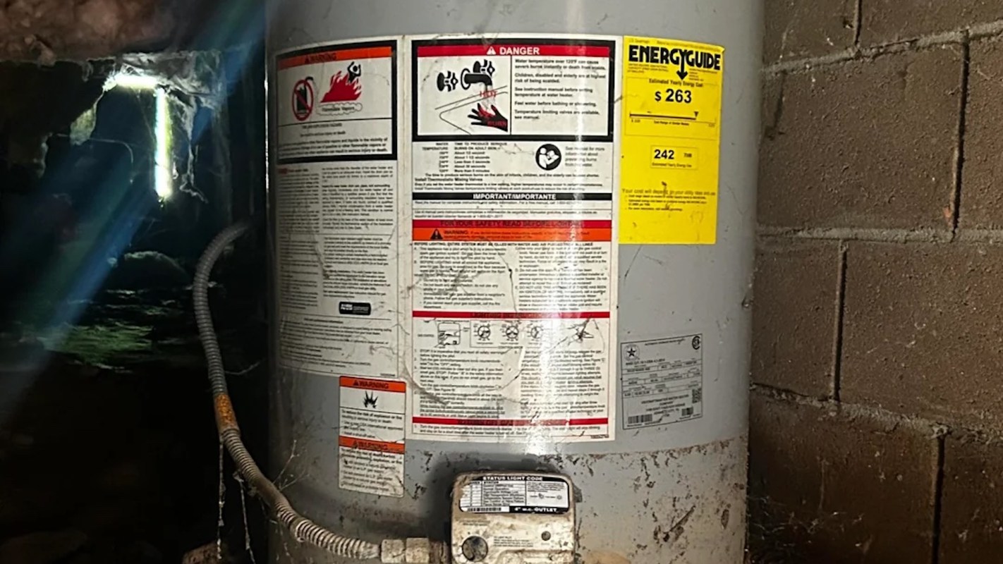 A water heater with warning labels and energy guide stickers set against a brick wall in a dimly lit space.