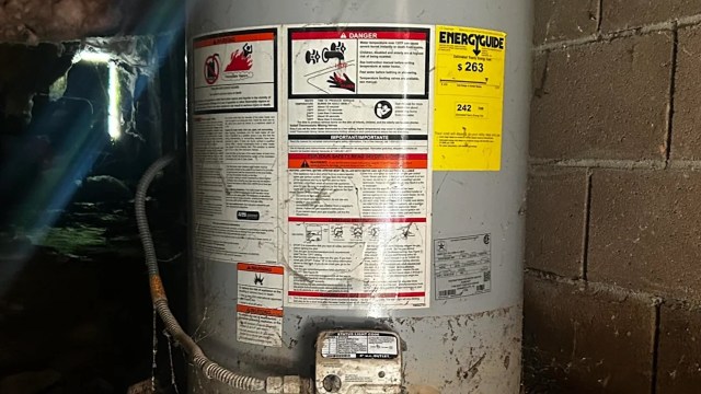 A water heater with warning labels and energy guide stickers set against a brick wall in a dimly lit space.