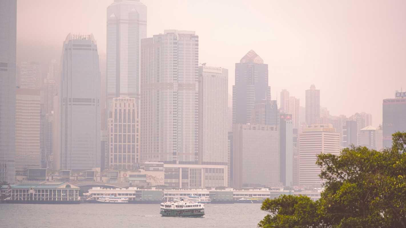 A ferry navigates through the harbor with a backdrop of hazy skyscrapers in a city skyline.