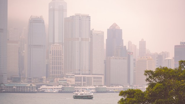 A ferry navigates through the harbor with a backdrop of hazy skyscrapers in a city skyline.
