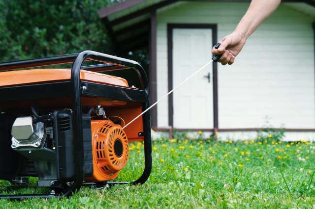 A person pulls the starter cord of a generator on grass near a white house.