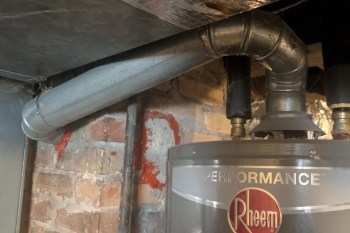 A close-up of a Rheem water heater with metal ducts and brick wall, showing cobwebs in the corner.