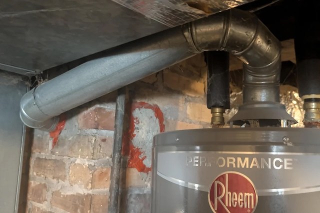 A close-up of a Rheem water heater with metal ducts and brick wall, showing cobwebs in the corner.