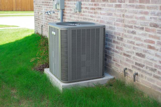 An air conditioning unit is positioned next to a brick wall, surrounded by lush green grass.