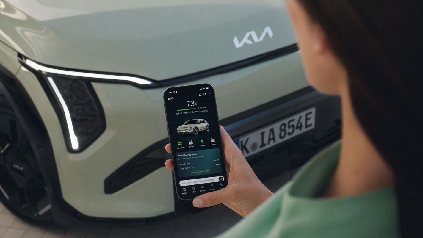 A person holds a smartphone displaying an electric vehicle control app in front of a Kia car.