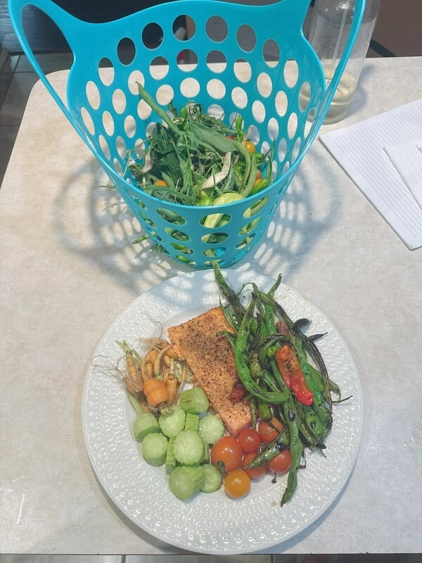 A dinner plate with fish and veggies next to a blue basket of the garden harvest. 