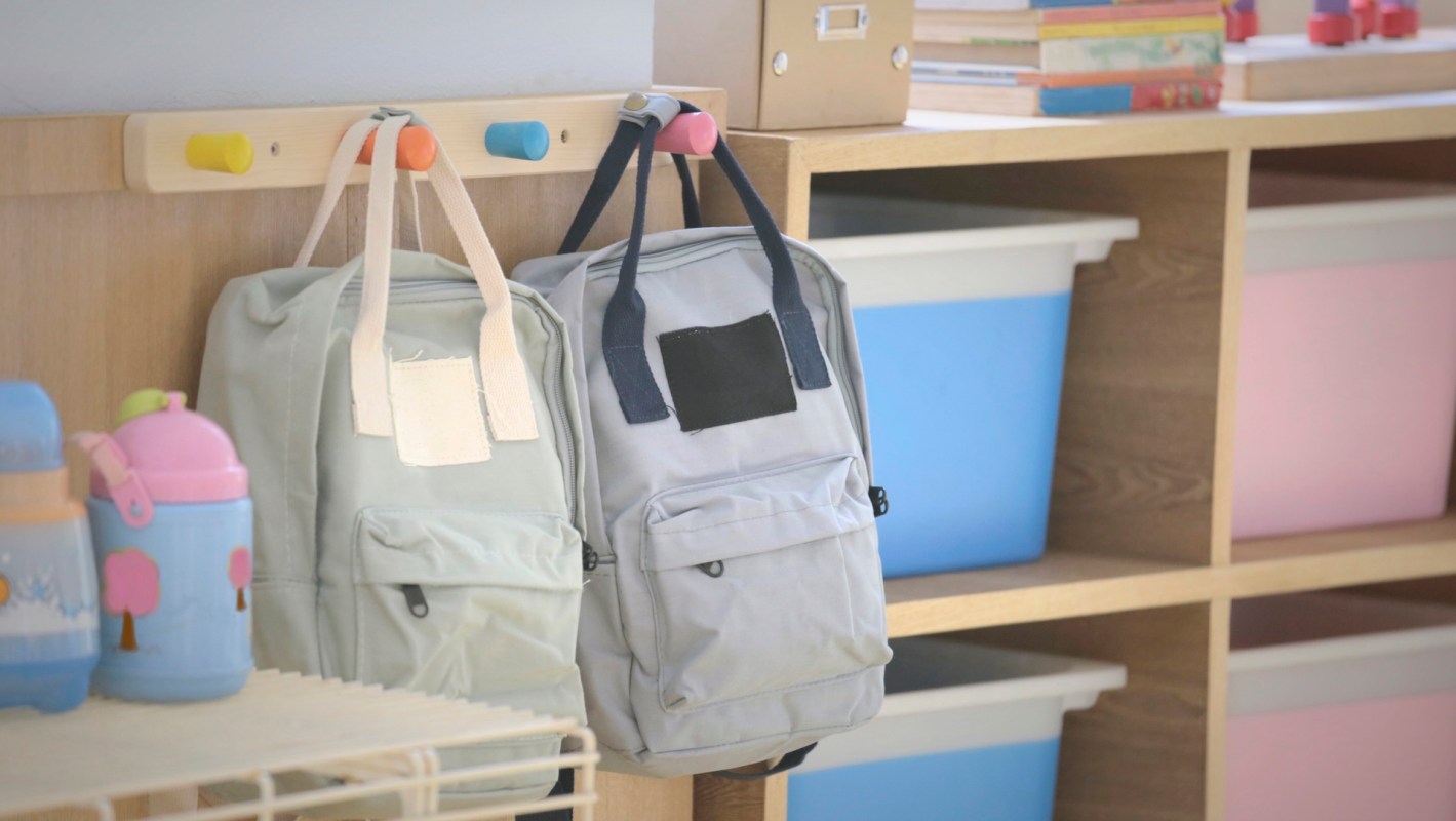 Two backpacks hang on a coat rack near colorful storage bins and children's books in a bright, organized room.