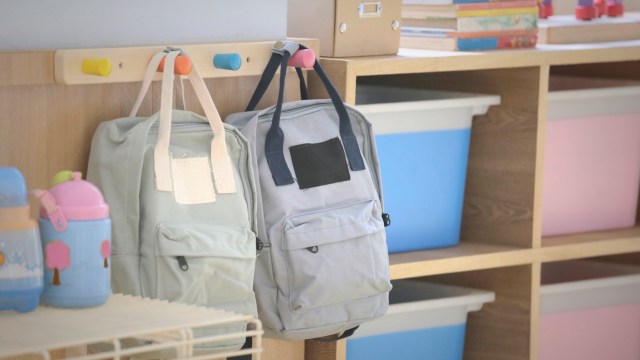Two backpacks hang on a coat rack near colorful storage bins and children's books in a bright, organized room.