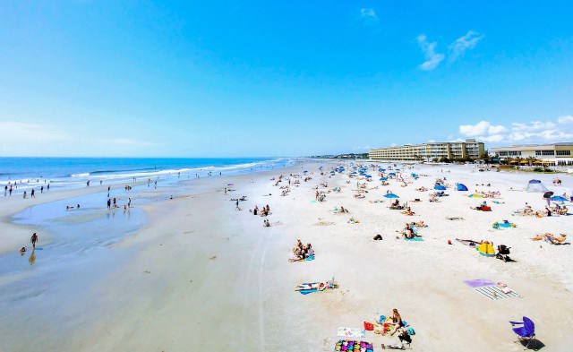 A busy beach scene with sunbathers, umbrellas, and people walking along the shore under a clear blue sky.