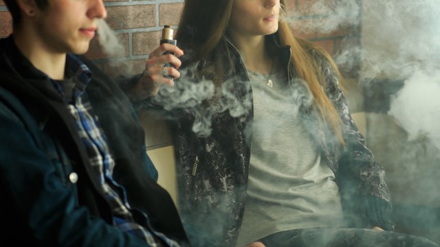 A young woman vapes while sitting beside a young man, surrounded by wisps of vapor and a brick wall backdrop.