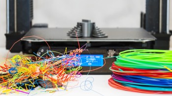 A 3D printer is in the background with a tangled mess of colorful filament in the foreground.