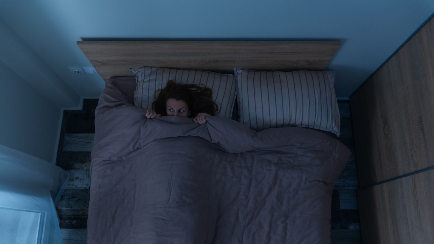 A woman sits in bed, peering out nervously from under her blanket in a dimly lit room.
