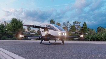 A futuristic flying vehicle with drone-like rotors is parked on a roadway amid trees and a blue sky.