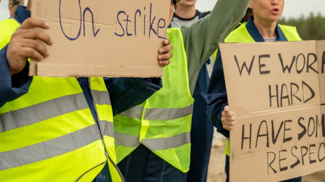 Workers wearing high-visibility vests hold cardboard signs protesting, one reads "On strike" and another "We work hard, have some respect."