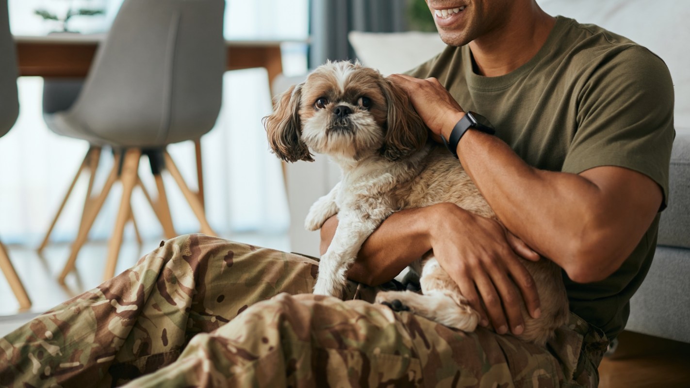 A person in military attire smiles while holding a small dog in a cozy indoor setting.